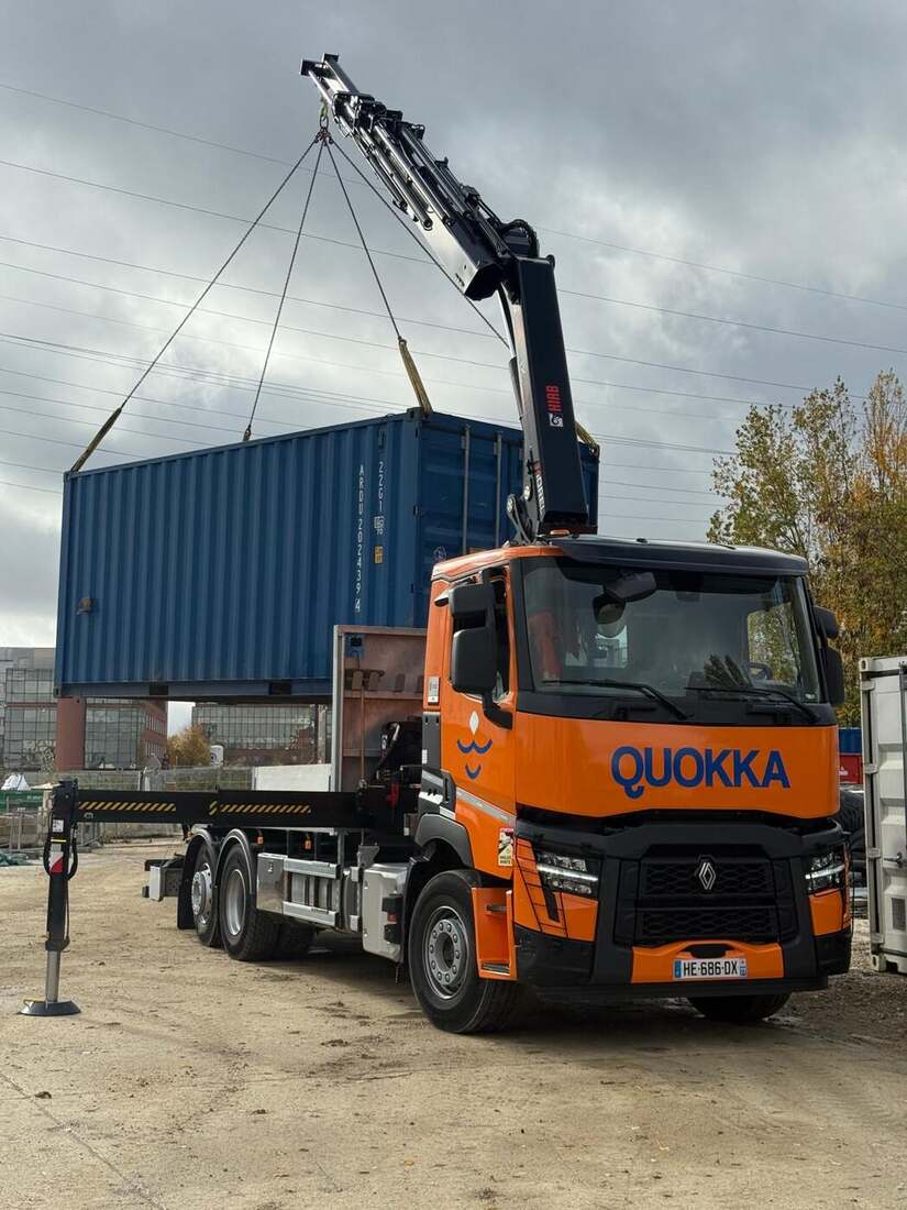 Camion-grue Quokka orange équipé d'une grue HIAB, en train de lever un conteneur maritime sur un chantier